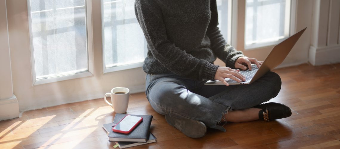 Close-up view of young female freelancer typing on laptop computer while sitting next to the windows with a cup of hot cocoa